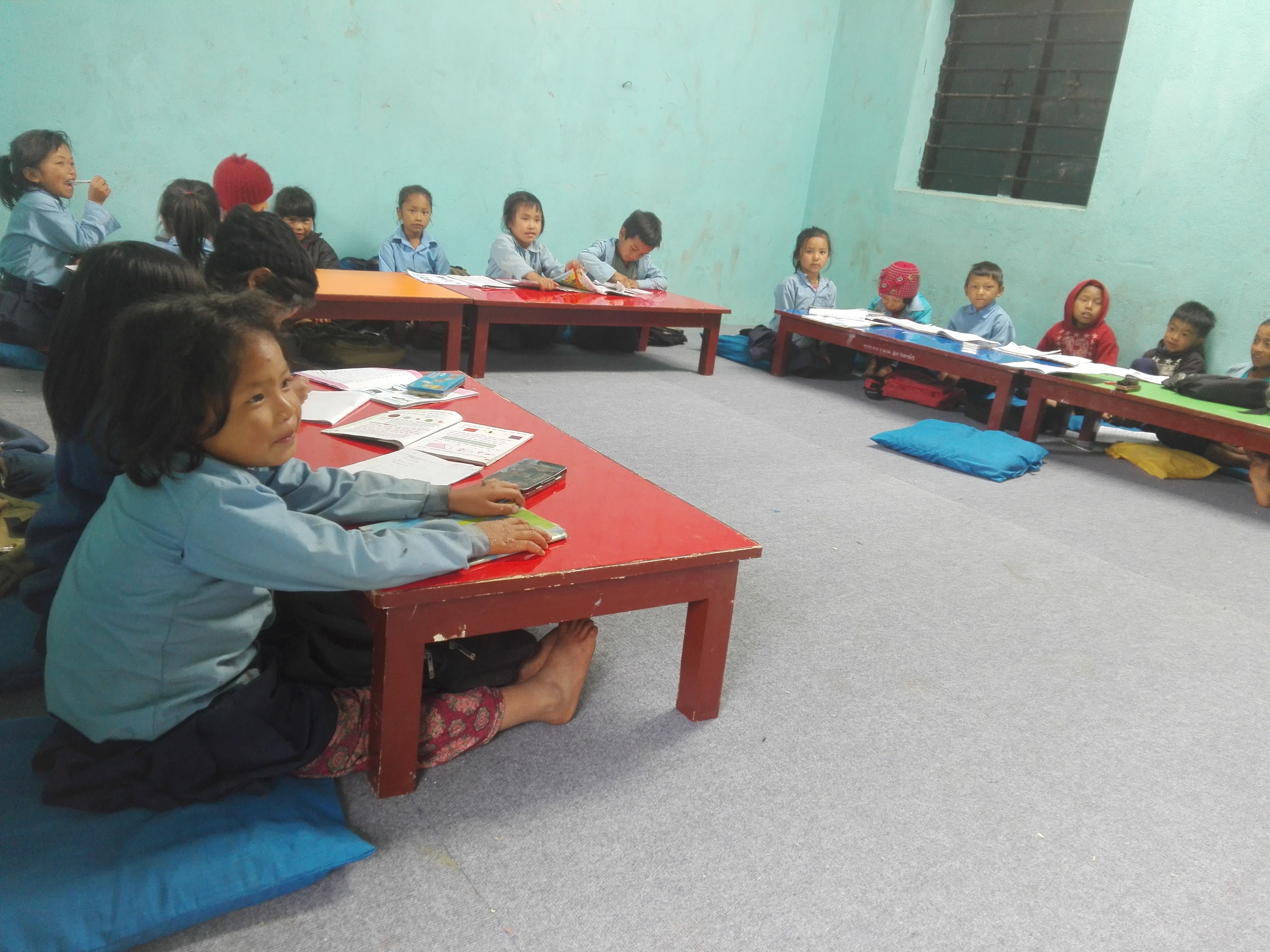 Bilde er fra et klasserpm med sementgulv. Barna sitter på puter rett på gulvet ved lave bord, veggene er helt nakne. Barna ser ut til å være ca 6-7 år gamle, og de bare føttene stikker fram under bordene rett på betonggulvet.